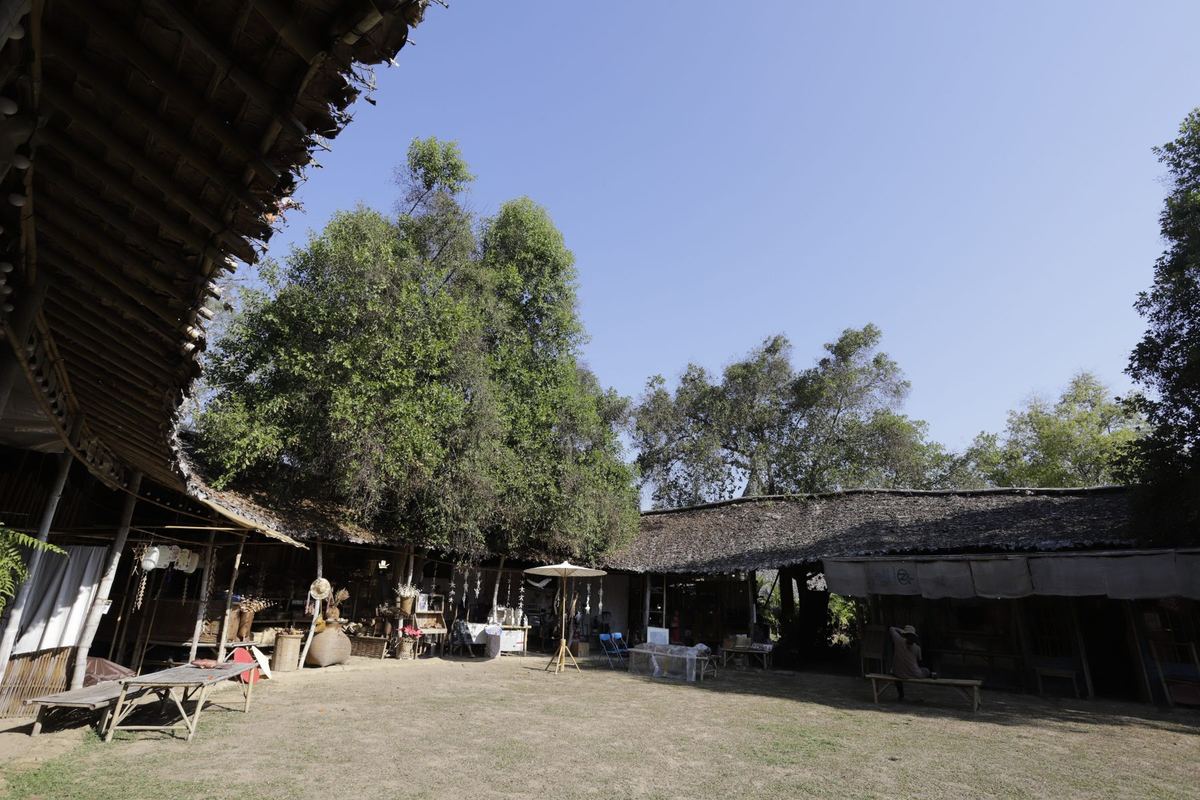 The courtyard under big trees, with thatched pavilions around a sandy yard